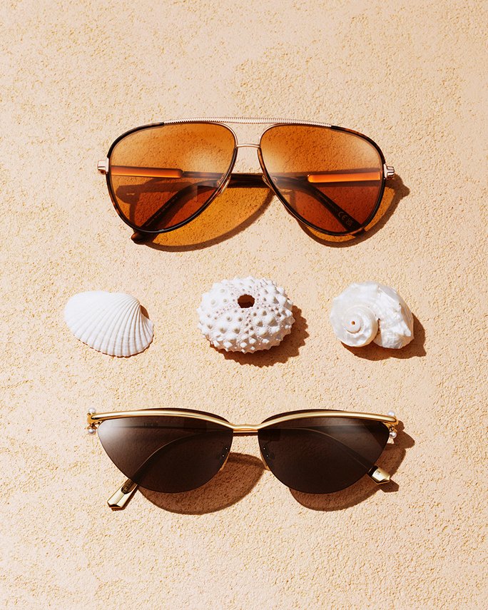Two pairs of sunglasses on a sandy beach, sea shells between them. Featured shops, the vacation shop.