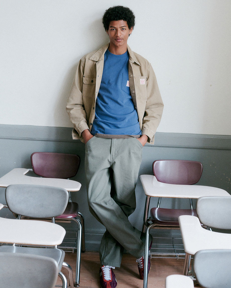A male college student standing in a classroom in a tan overshirt, blue tee & green pants. Top picks for the new year, men.