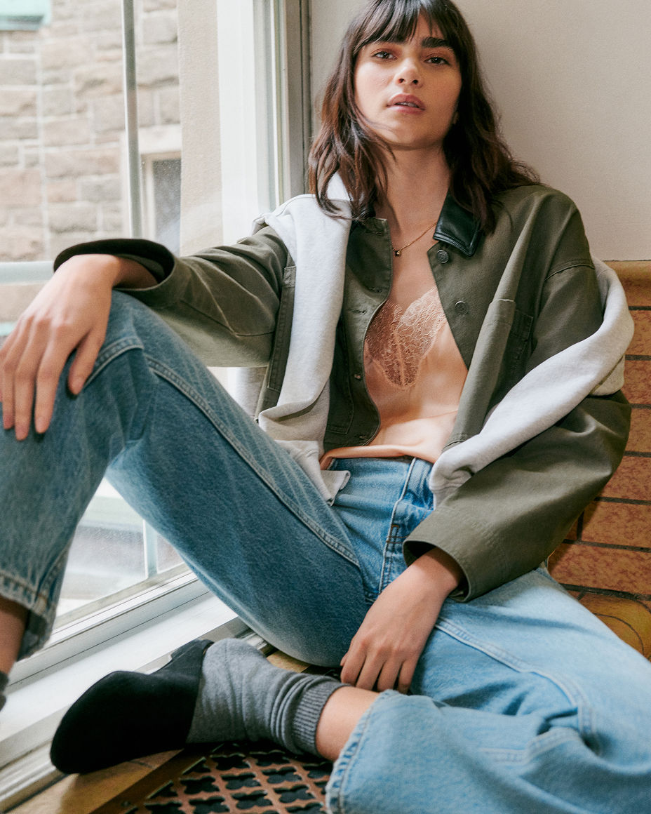 A college student sitting casually on the floor in a green barn jacket, pink lacy top & jeans. Featured shops, back to campus.