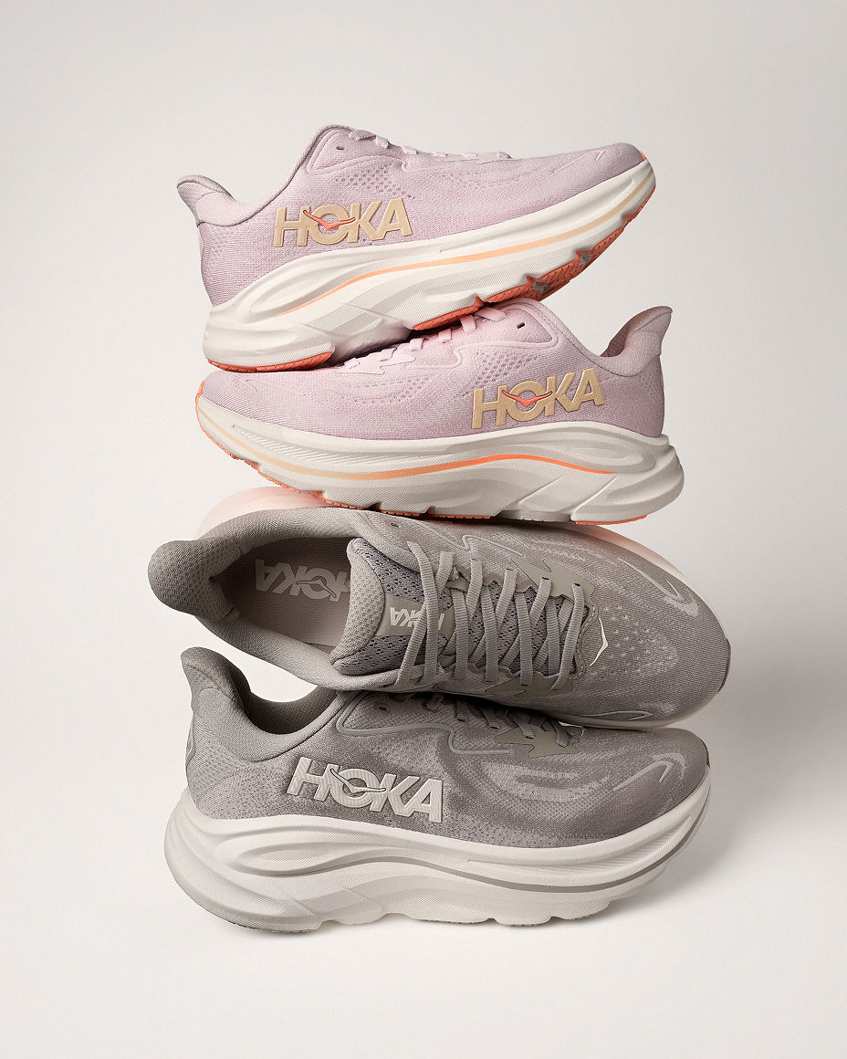 A pink pair of Hoka sneakers balanced on a pair of gray Hoka sneakers. Featured shops, the sneakers guide.