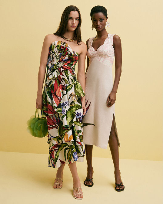 Two women in Farm Rio summer dresses, one a strapless tropical plant print, the other a white sleeveless. Shop dresses.