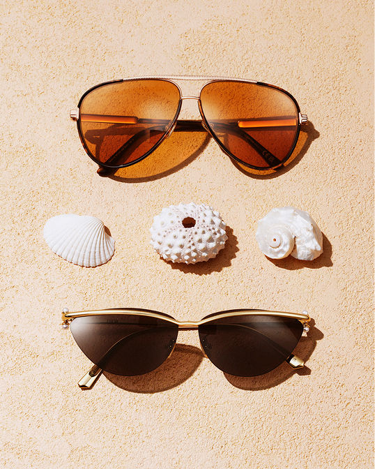 2 sunglasses on a beach, three seashells lined up between them. Shop sunglasses.