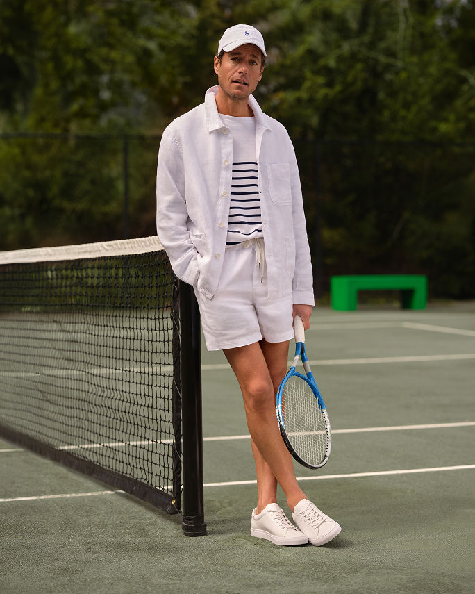 A man on a tennis court in an all white tennis outfit. Get outdoors. Shop mens activewear.