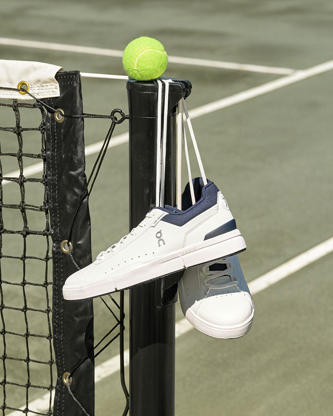 A pair of white On tennis sneakers hanging from the end of a tennis net. Get outdoors. Shop mens sneakers.