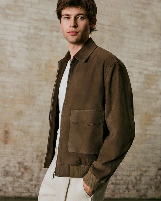 A man in a brown suede jacket over a white top and stone colored pants. Shop men.