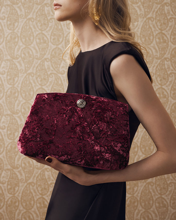 A woman holding a large raspberry colored crushed velvet clutch. Shop clutches.