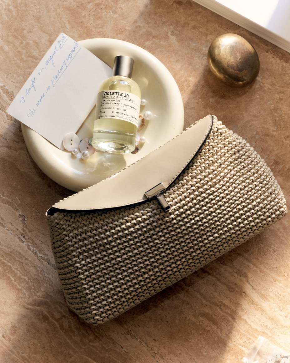 Brown textured clutch next to a bowl with a bottle of perfume & odds & ends.