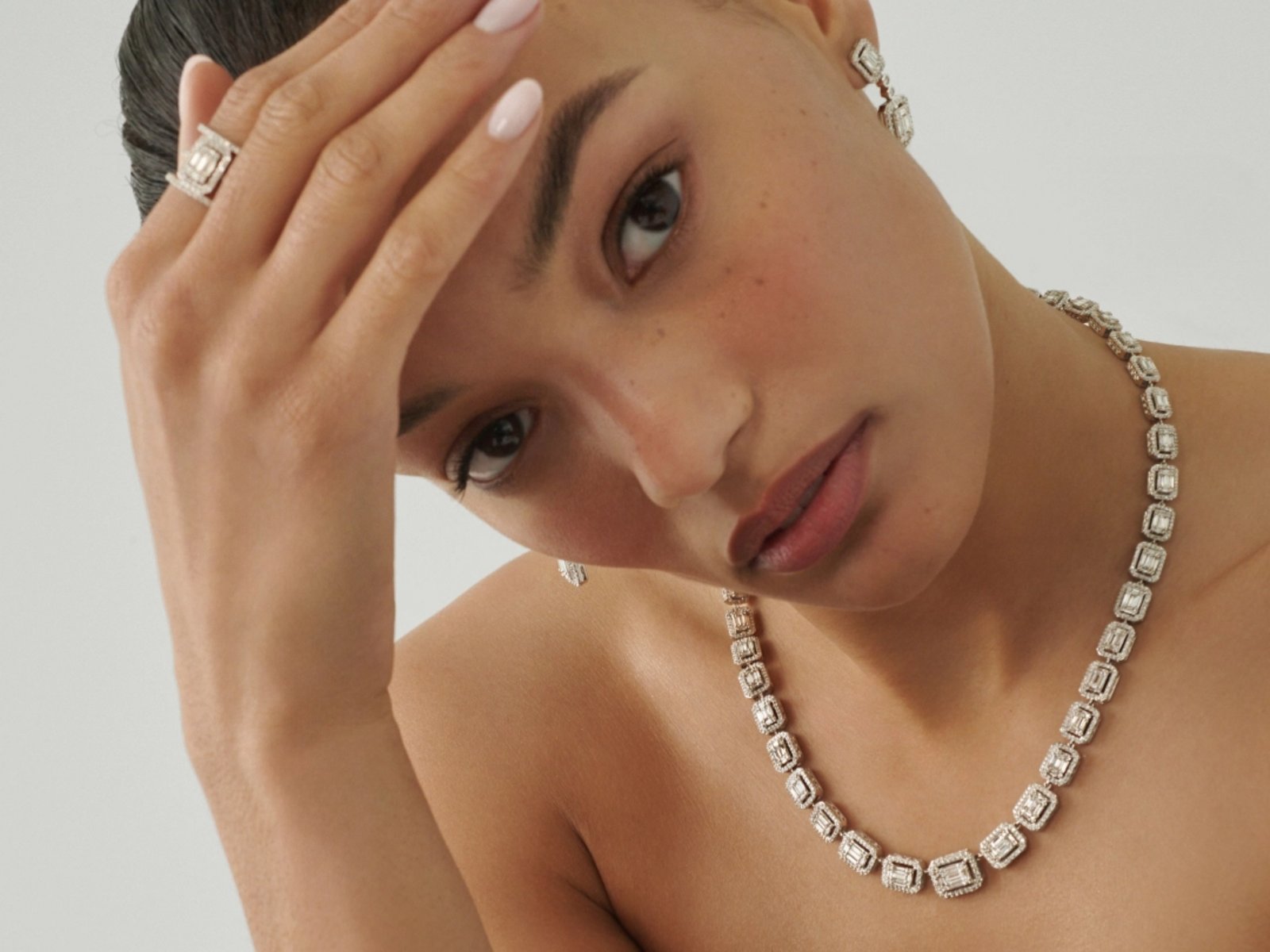 1 photo, a closeup of a female model, one hand on her forehead, wearing a sparkling Bloomingdale's Fine Collection mosaic diamond set, including drop earrings, a tennis necklace and bracelet, and a ring. 