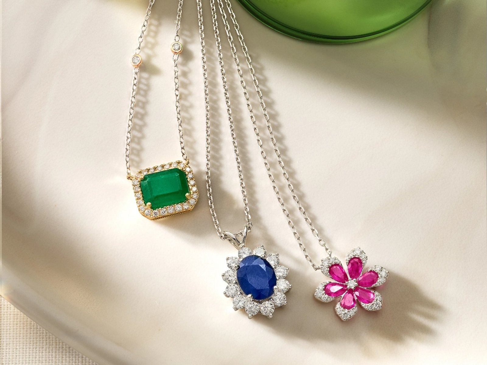 1 photo, of 3 pendant necklaces on a marble background, a square cut emerald lined with diamonds, an oval sapphire lined with flower shaped diamonds, and pink gemstones and diamonds shaped like flower petals.