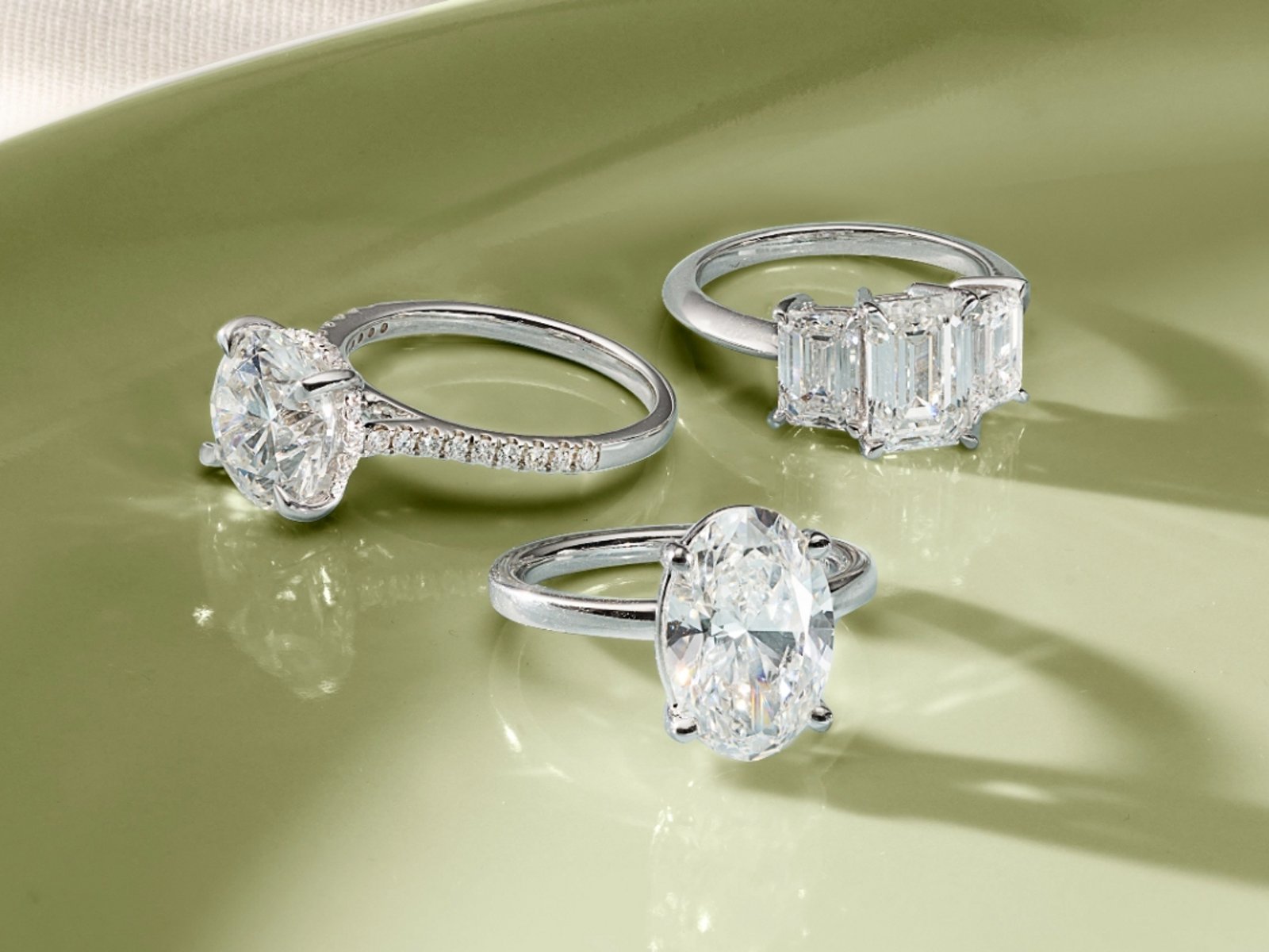 1 photo, of 3 sparkling diamond solitaire rings with white gold bands, in baguette, round, and oval cuts, on a shiny green surface.