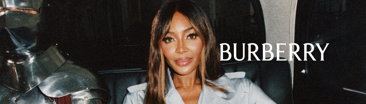 Closeup of model Naomi Campbell in a light blue Burberry trench coat sitting in a car next to a man in a full suit of silver armor.