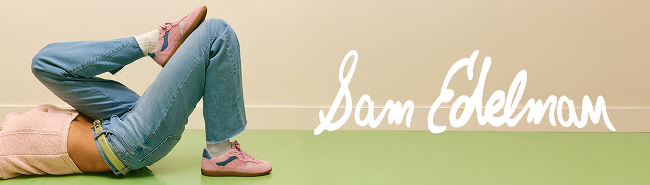 1 photo, of a female model from the waist down, lying on their back, one leg propped on the other leg, wearing a pink top, blue jeans, and pink Sam Edelman sneakers.