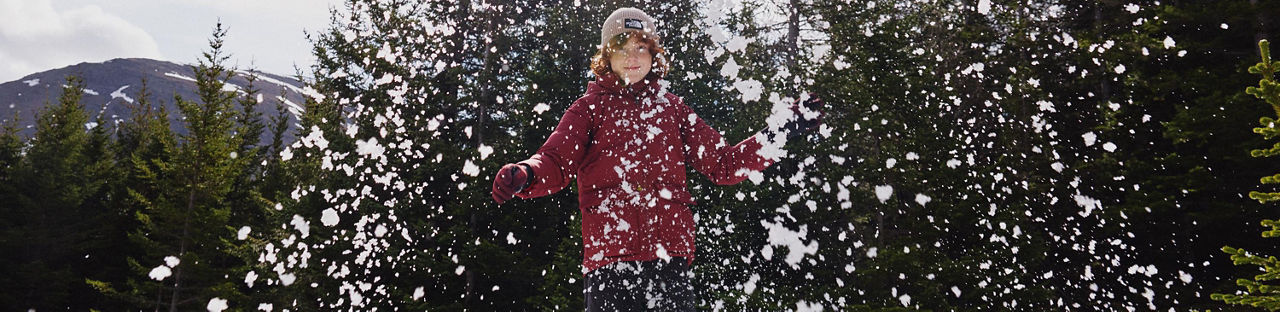 A boy in a North Face red puffer jacket throwing snow, evergreens behind him.