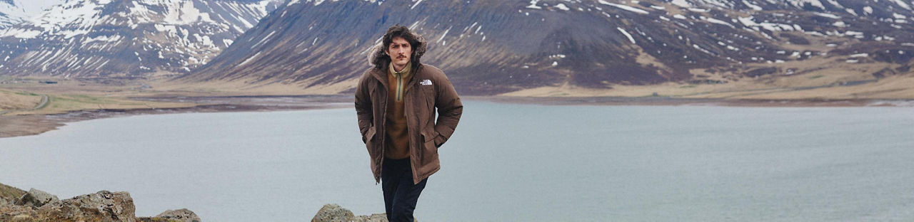 A man in a North Face brown parka standing in front of a mountain lake.