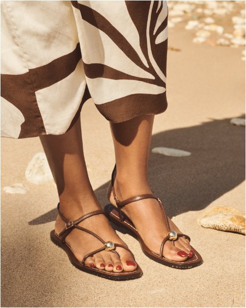 A womans feet in brown strappy flat sandals with a pearl embellishment.