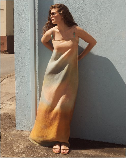 A woman leaning against a wall in a painterly pastel peach and gray sleeveless maxi dress and sunglasses.